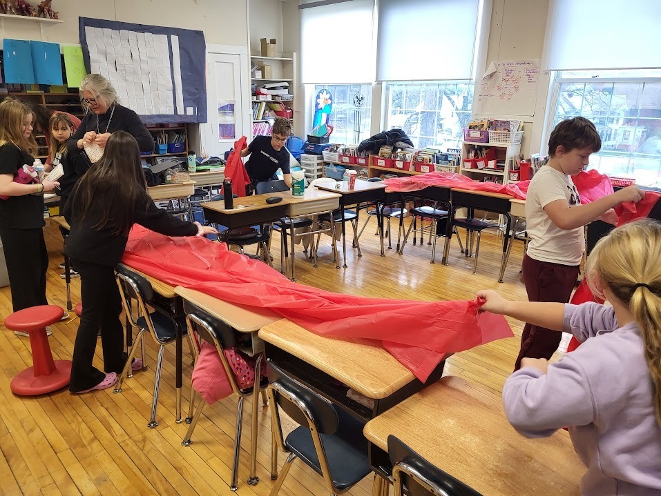 Students and a teacher work together in a classroom to lay out long sheets of red tablecloth material across several desks in preparation for a meal.