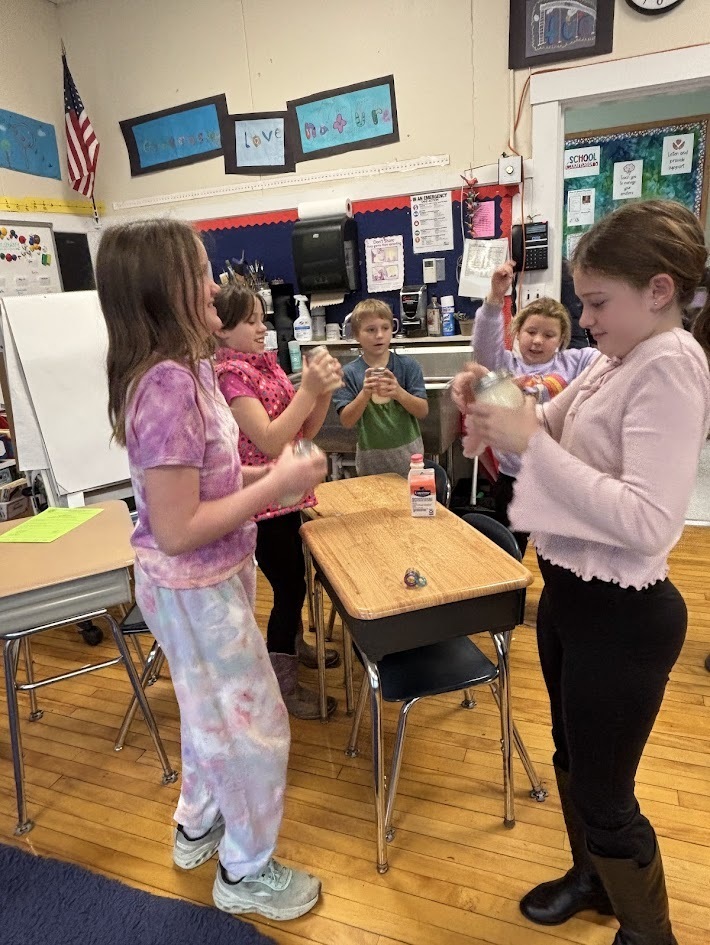 A group of elementary students stands around desks shaking jars, likely making butter. They smile and laugh in a bright classroom decorated with posters and student artwork.
