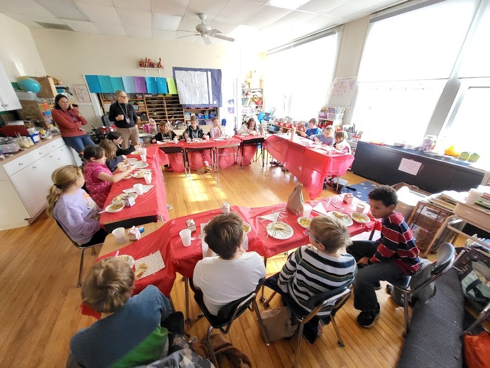 A classroom of students sits around a large U-shaped arrangement of desks covered with red tablecloths, eating a meal together. Teachers stand at the back of the room.