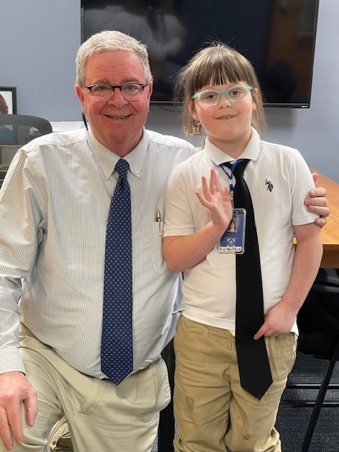 A young student dressed in a white shirt and tie stands proudly with an adult man, also in a shirt and tie. They smile in what appears to be an office or conference room, the student wearing ID cards around their neck.