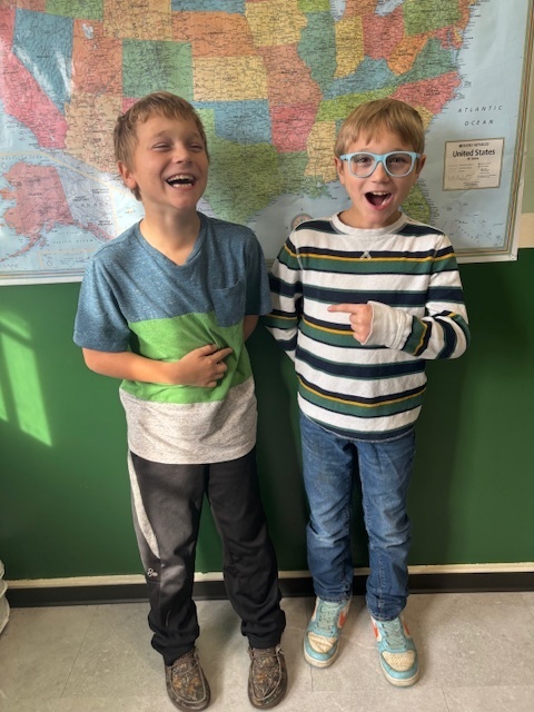 Two young boys stand in front of a classroom map of the United States. One boy wears blue glasses and a striped sweater and is pointing excitedly; the other boy laughs, wearing a blue-and-green color-block shirt.