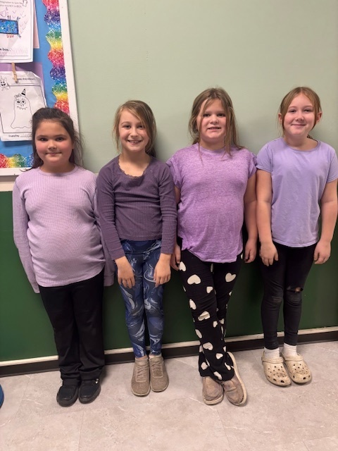 Four girls stand in a row against a light green wall, all dressed in purple tops. They smile as they pose together.