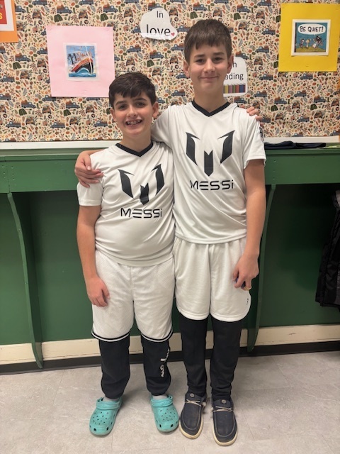 Two boys stand side-by-side wearing matching white “Messi” soccer uniforms. One rests his arm on the other’s shoulder, and both smile proudly.