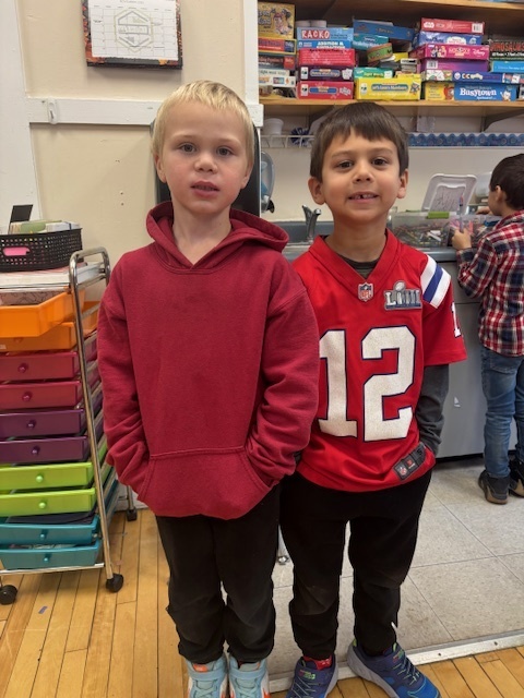 Two boys stand in a classroom; one wears a red Patriots jersey and the other a red hoodie. They smile as they pose in front of colorful classroom materials.