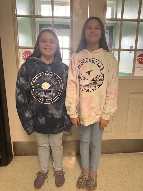 Two students stand side-by-side in a hallway, both wearing “Willoughby Lake, Vermont” hoodies—one in a light tie-dye style and the other in a blue-and-gray tie-dye. They smile at the camera with a set of windows behind them.