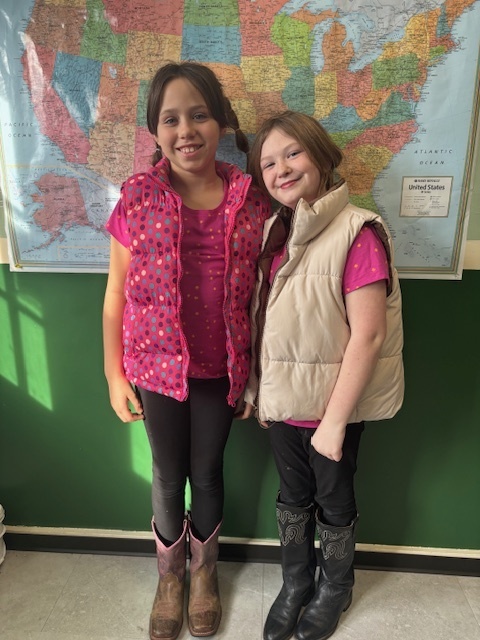 Two girls stand in front of a map, dressed in pink-themed outfits. One wears a beige vest and pink shirt, and the other wears a pink polka-dot vest. Both wear boots and smile at the camera.