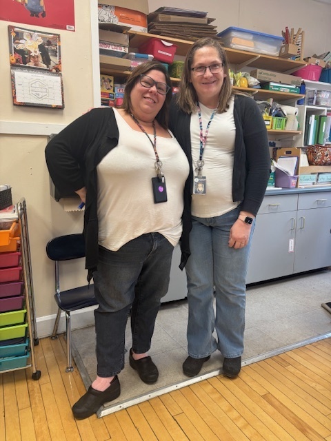 Two teachers stand in a classroom, wearing unintentionally matching outfits: white tops, black cardigans, and jeans. They smile warmly.