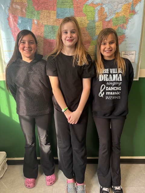Three girls stand in front of a large U.S. map, all dressed in black outfits. They smile brightly, with sunlight shining on them from the side.