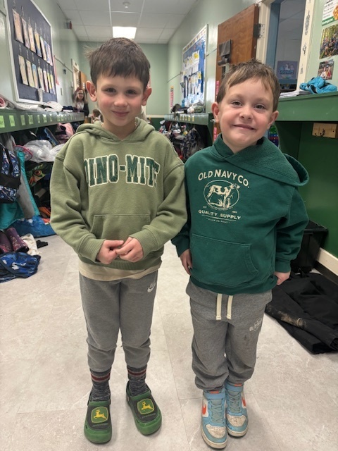 Two boys stand together in a hallway, wearing green hoodies—one with an Old Navy logo and the other with a John Deere design. They smile at the camera.