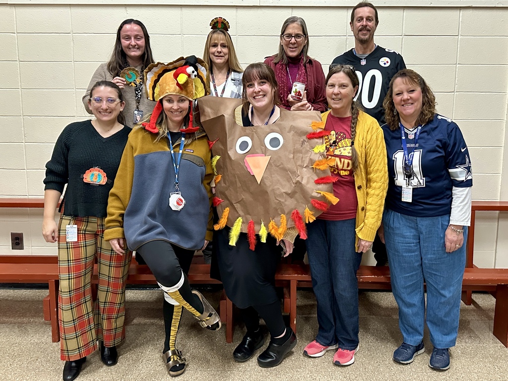 A group of nine CES staff members pose together indoors, all dressed to match a Thanksgiving theme. Some wear turkey hats, fall colors, or shirts with turkey graphics. One staff member is dressed in a large handmade turkey costume with colorful feathers. The group stands smiling in front of a light-colored brick wall and red bench seating.
