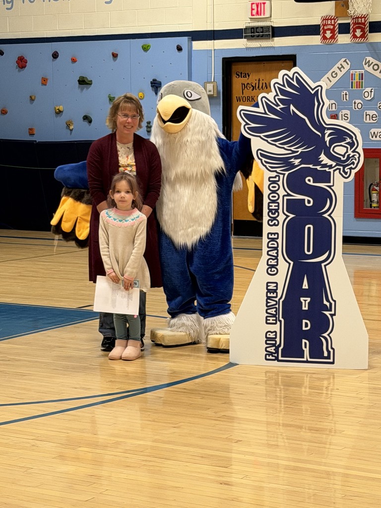 One adults, one child holding certificate, and a school mascot stand together in a gymnasium for a photo.