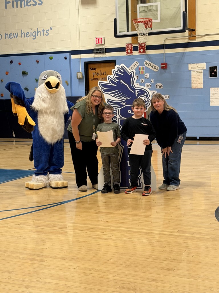 Two adults, two children holding certificates, and a school mascot stand together in a gymnasium for a photo.