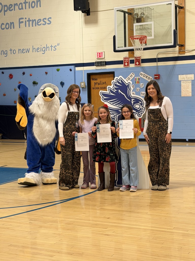 Two adults, three children holding certificates, and a school mascot stand together in a gymnasium for a photo.