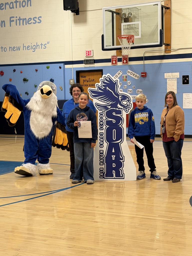 Two adults, two children holding certificates, and a school mascot stand together in a gymnasium for a photo.