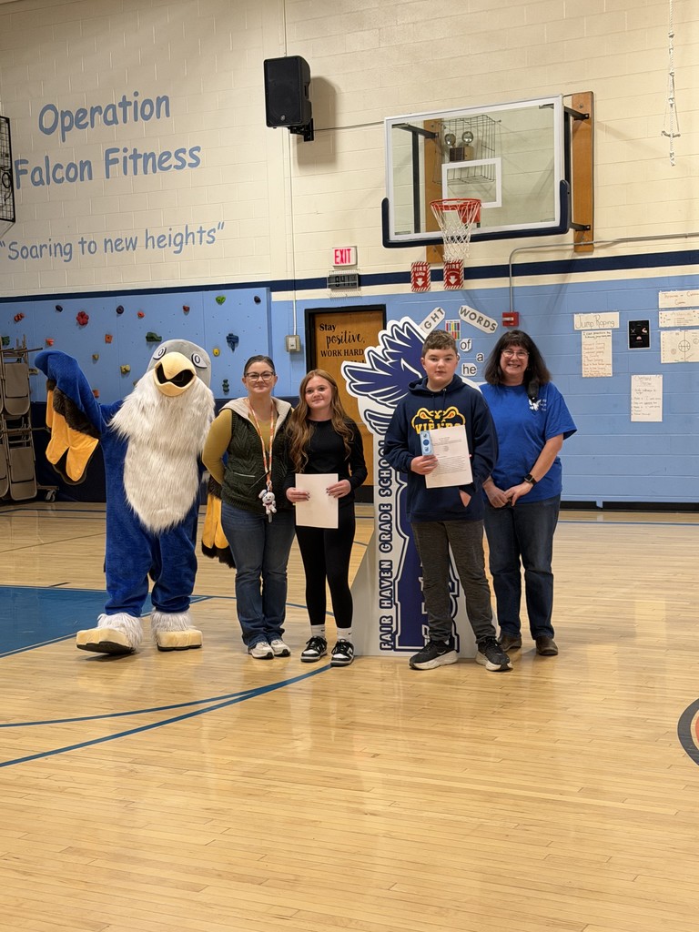 Two adults, two children holding certificates, and a school mascot stand together in a gymnasium for a photo.