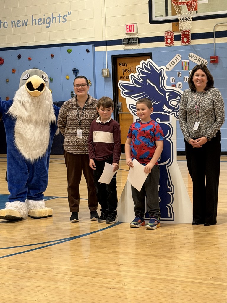 Two adults, two children holding certificates, and a school mascot stand together in a gymnasium for a photo.