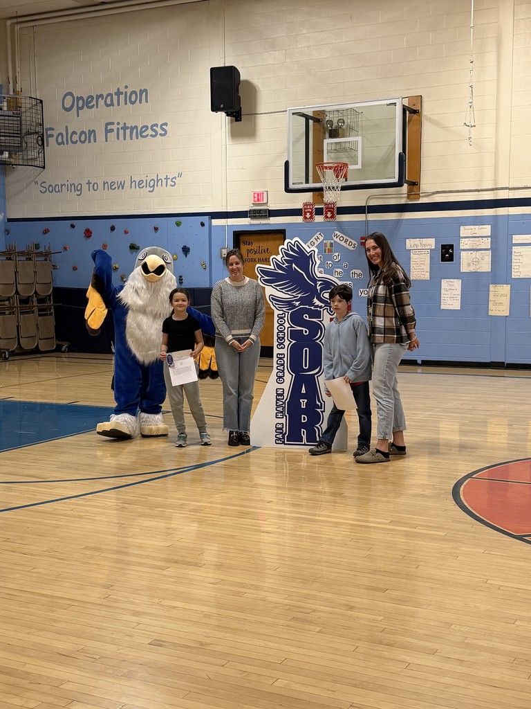 Two adults, two children holding certificates, and a school mascot stand together in a gymnasium for a photo.