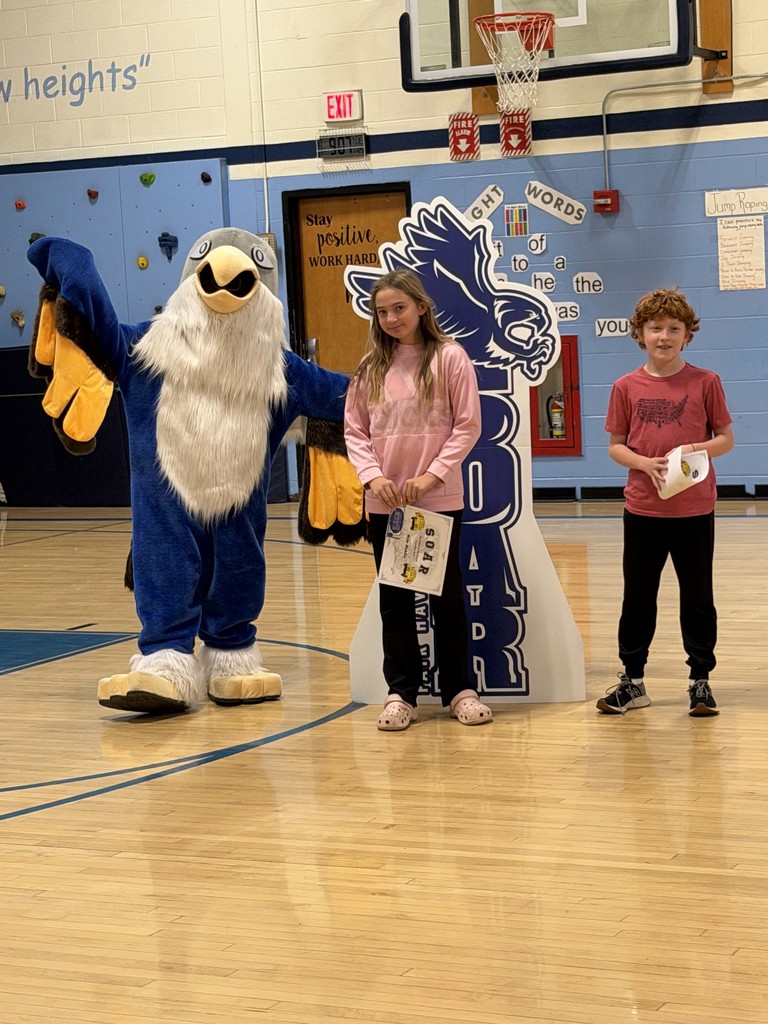 Two children holding certificates, and a school mascot stand together in a gymnasium for a photo.