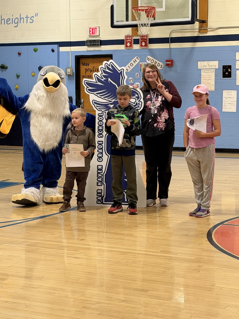 One adult, three children holding certificates, and a school mascot stand together in a gymnasium for a photo.