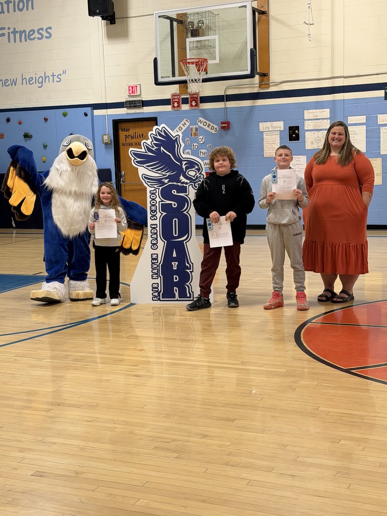 One adult, three children holding certificates, and a school mascot stand together in a gymnasium for a photo.