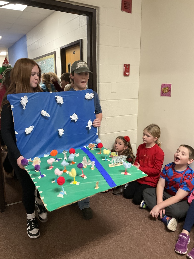  Two older students present a diorama made of a blue background with cotton balls for clouds and a green base decorated with small, colorful pom-pom trees to a group of three younger children sitting on the floor in a school hallway.