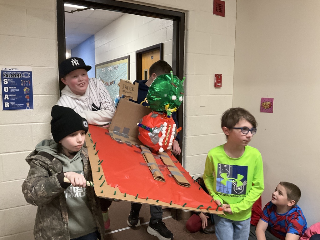 Five young students hold a large, angled cardboard project decorated with red material and what appears to be a green balloon or structure at the top, standing in a hallway doorway. Two boys are supporting the structure, while another wears glasses and a bright green shirt.