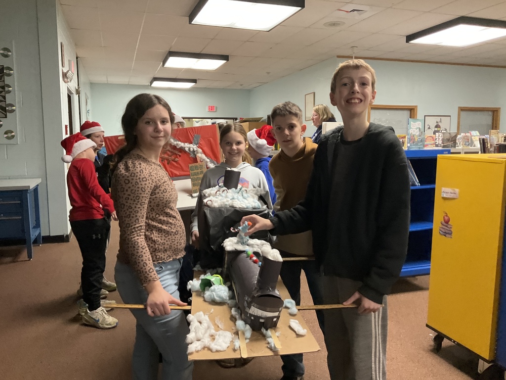 Group of four middle school-aged students, smiling and posing in a hallway with a large, decorated, boat-like project covered in cotton "snow" on a cardboard base. Other students wearing Santa hats are visible in the background.
