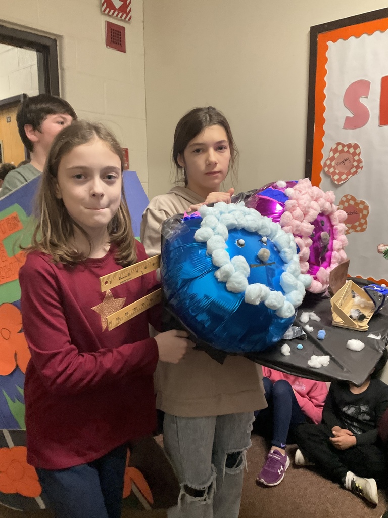Two students hold a colorful balloon-based craft decorated with cotton “snow” in a school hallway, with classmates seated in the background.