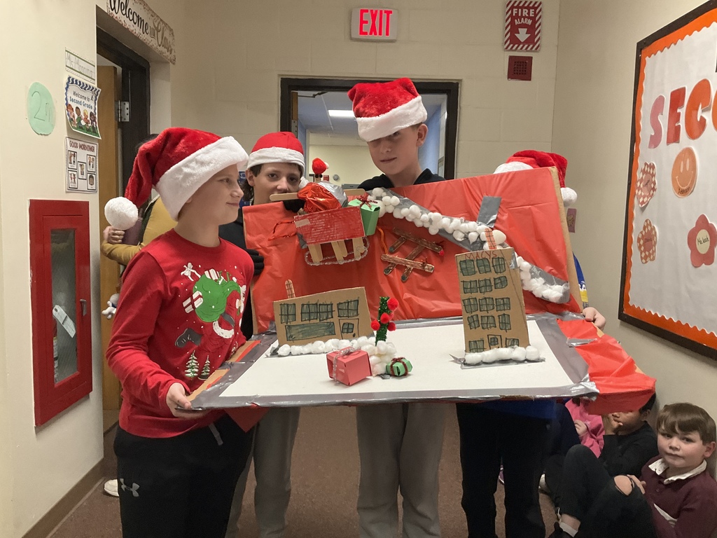 Students wearing Santa hats hold a large holiday-themed cardboard display featuring buildings, cotton “snow,” and festive decorations in a school hallway.