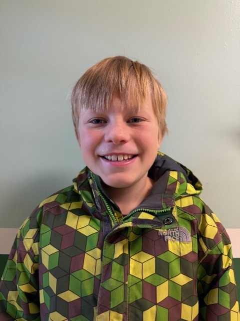 A young boy with light brown hair smiles at the camera while wearing a green, yellow, and brown geometric-patterned jacket.