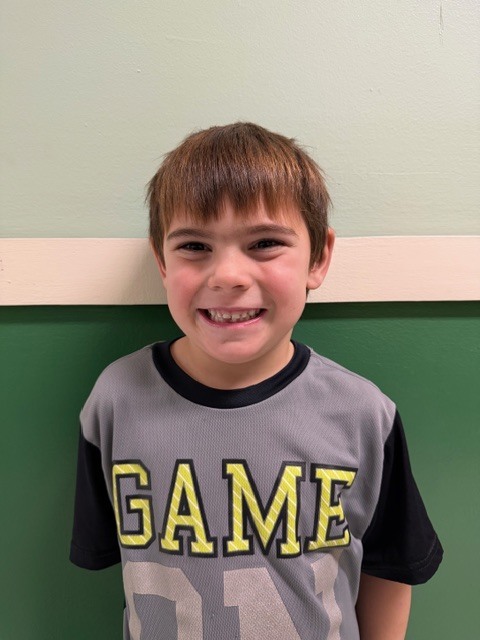 A young boy with short brown hair smiles while wearing a gray shirt with the word “GAME” in large yellow and white letters.