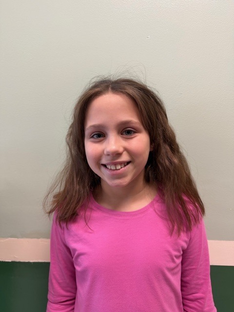A girl with shoulder-length brown hair smiles at the camera while wearing a bright pink long-sleeve shirt.