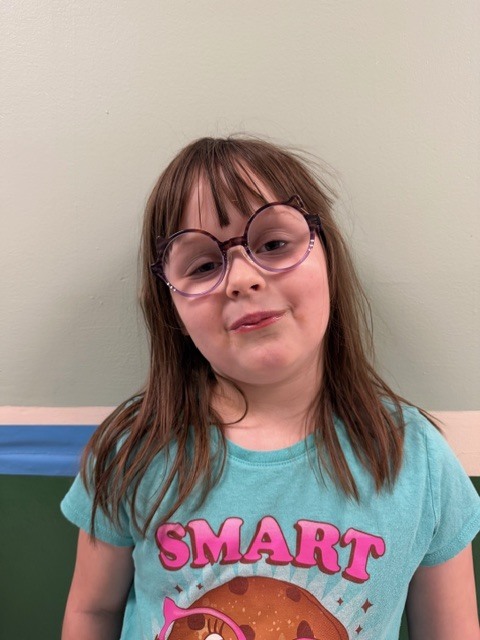 A young girl with long brown hair and purple glasses smiles softly while wearing a teal shirt with colorful graphics.