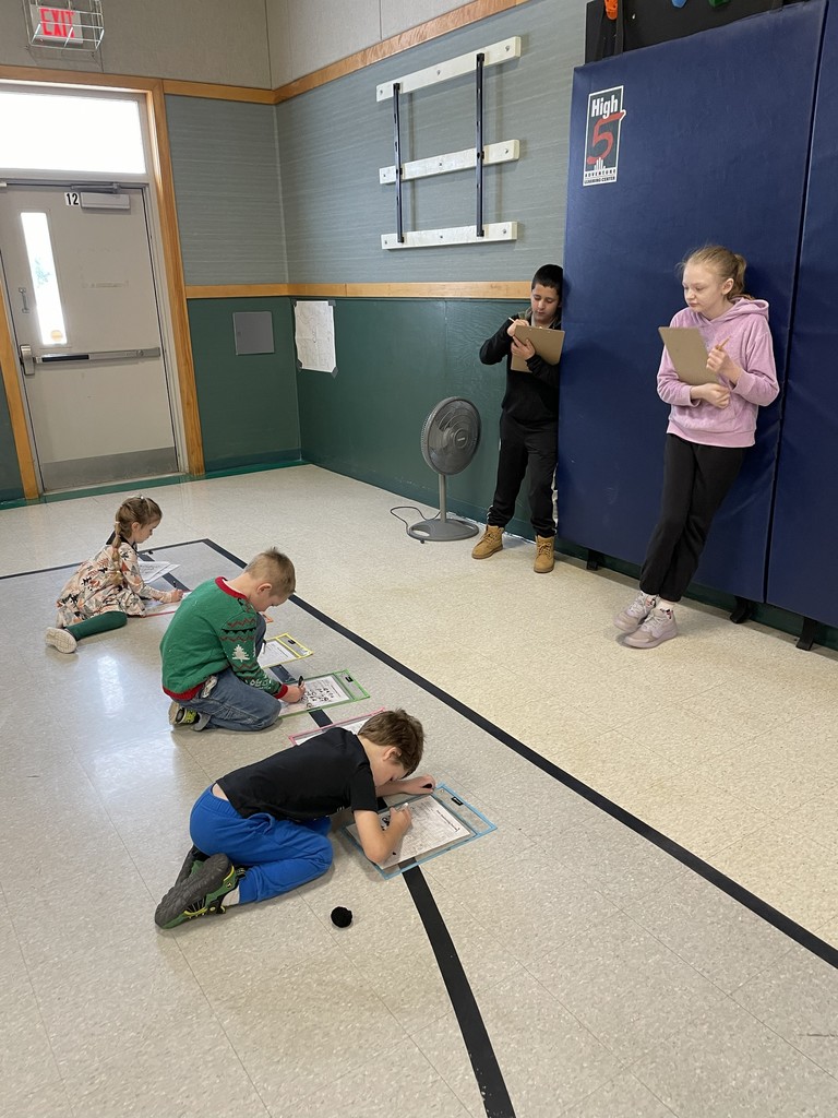 Six students are in a large room, likely a gymnasium, participating in an activity. Four younger children are sitting or lying on the floor, writing on clipboards. Two older children are standing against a blue padded wall, also writing on clipboards.  