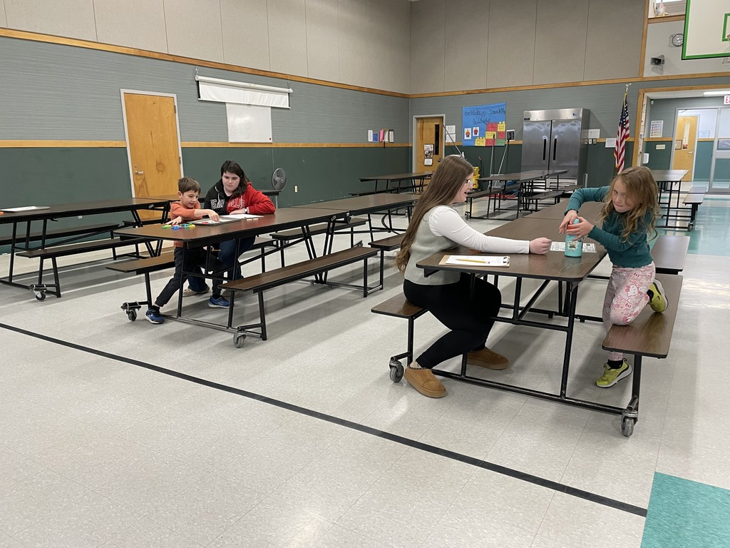  A large, brightly lit cafeteria or multi-purpose room with several students and adults seated at long folding tables. Two adults are helping four younger children with an activity that involves writing and small manipulatives on the tables.