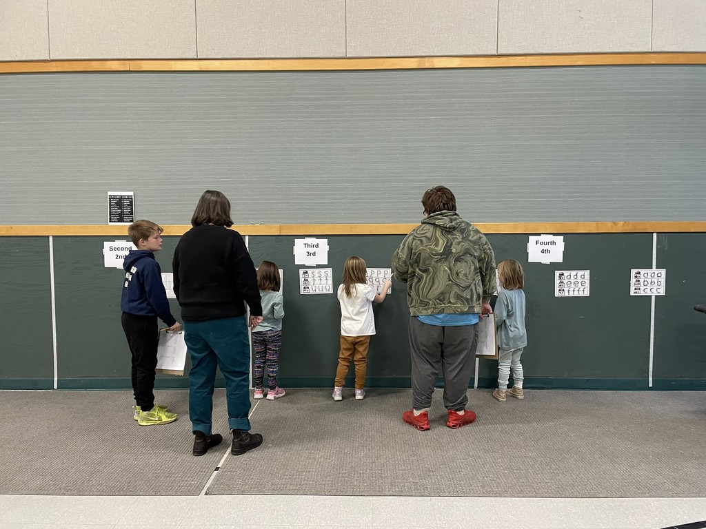  A group of six people, including adults and children, stand facing a green wall that has large printouts attached to it, labeled by grade levels (Second, Third, Fourth). The children are tracing or marking letters on the printouts, while the adults observe.