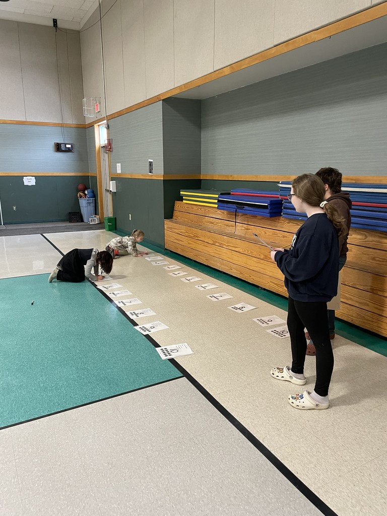 n a gymnasium, two young girls are crawling along a long line of papers taped to the floor that have printed words or numbers on them. Two older students stand nearby, observing the activity and holding clipboards.
