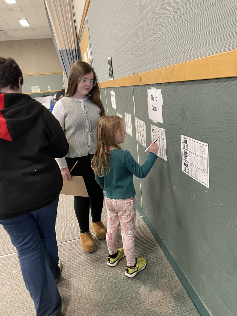  A close-up view of a young girl in a green top and patterned pants tracing letters on a printed worksheet attached to a wall, while two older individuals (an adult and an older student) stand next to her, watching and holding a clipboard. The section of the wall is labeled "Third 3rd". 