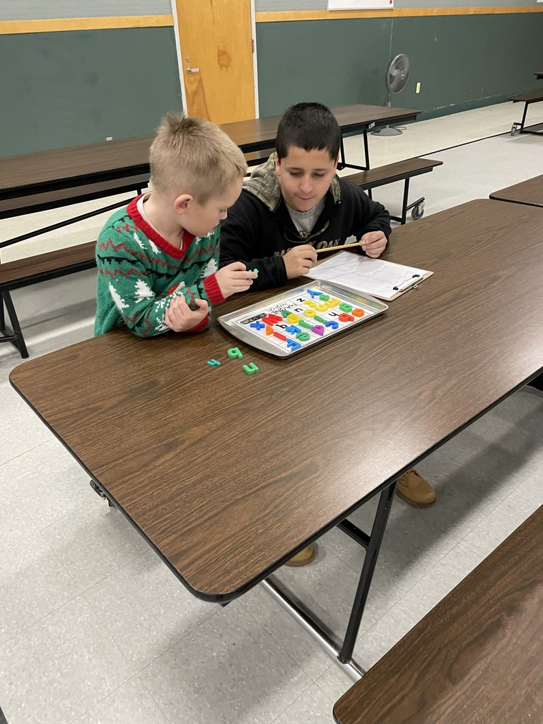 Two boys, one in a green holiday-themed sweater, are sitting across a brown cafeteria table from each other. They are engaged in an educational activity using colorful, magnetic letters laid out on a small metal tray. The boy on the right is writing on a clipboard. 