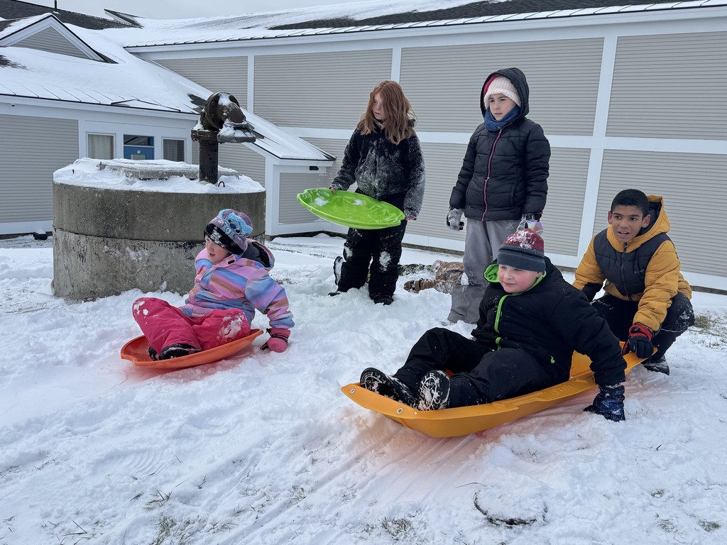 Five children standing and sitting around plastic sleds (orange, green, and red) on a snow-covered area next to a building. The children are bundled up in winter gear and smiling.  IMG_9589.jpg: Two girls sitting on a red plastic toboggan-style sled in the snow near a large concrete structure. The girl in front, wearing a pink snowsuit, is holding the rope and smiling, while the girl behind is sitting with her.
