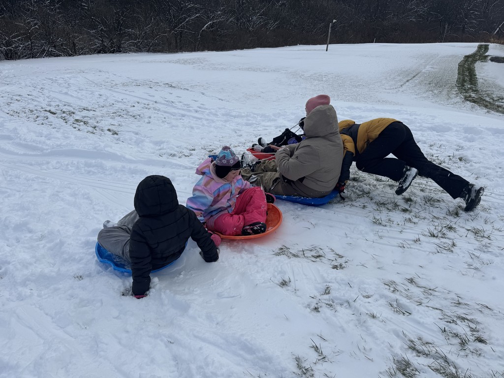 A group of children gathered at the bottom of a snowy hill. Three are sitting on plastic sleds (one in black, one in pink/multi-color, one in beige), while a fourth person in a yellow and black jacket is leaning forward on the ground next to them.  