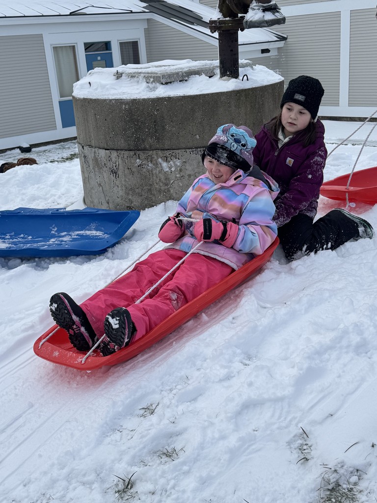 Two girls sitting on a red plastic toboggan-style sled in the snow near a large concrete structure. The girl in front, wearing a pink snowsuit, is holding the rope and smiling, while the girl behind is sitting with her.