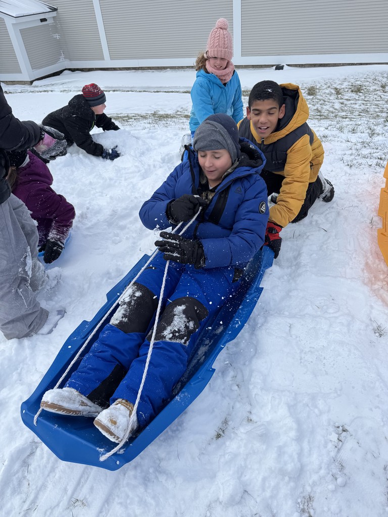  A group of children playing in the snow. A boy in a blue snowsuit and grey hat is sitting on a blue sled, holding the pull rope, while another boy in a yellow jacket and a girl in a pink hat kneel beside him. 