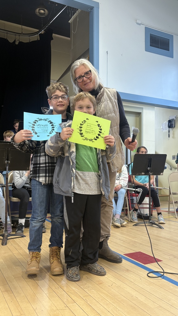 A teacher stands with two boys holding blue and yellow certificates during a school assembly.