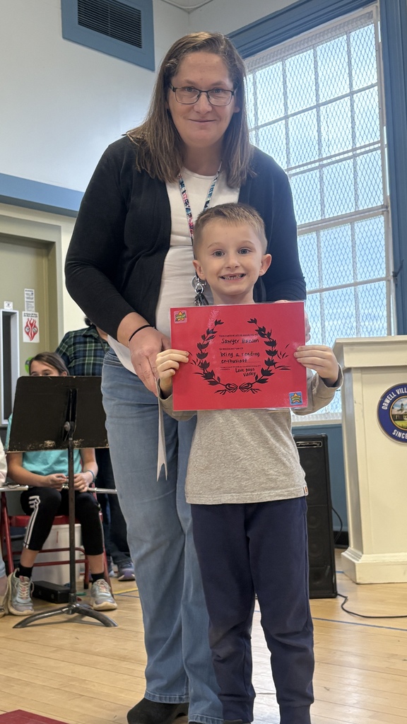 A teacher stands with a smiling boy holding a red award certificate in the school gym.