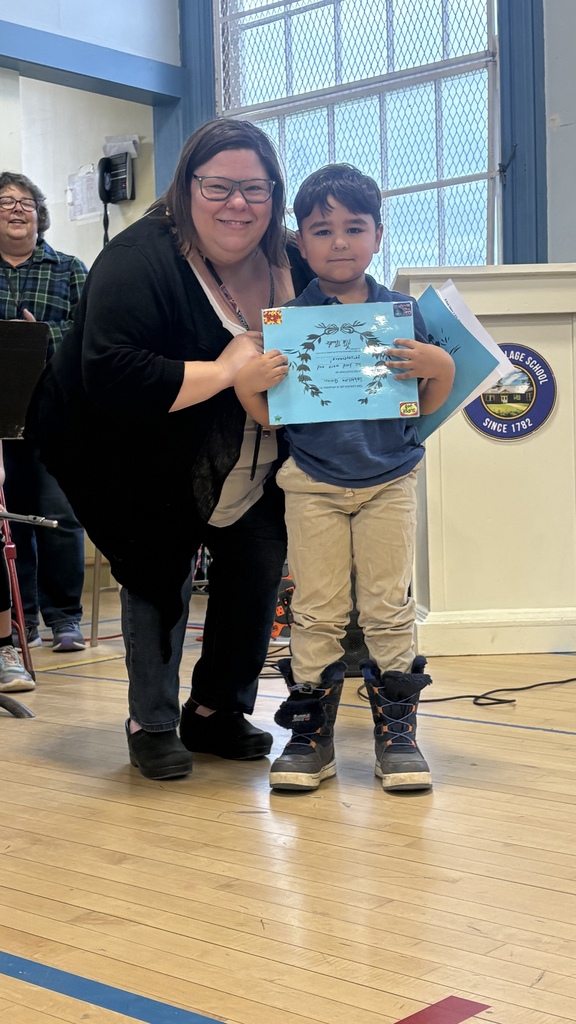 A teacher kneels beside a young boy holding a blue award certificate during a school assembly.