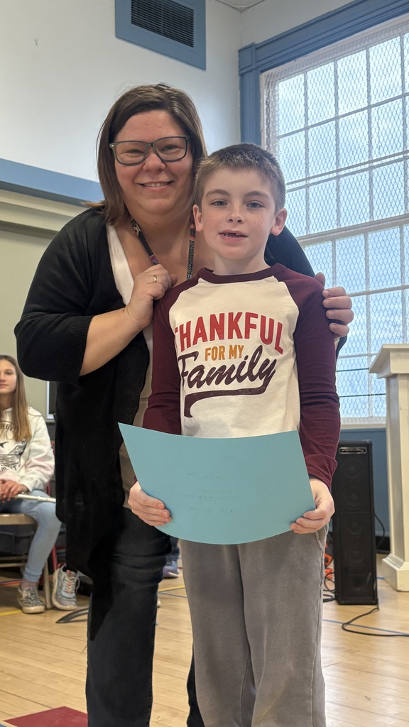 A teacher poses with a boy wearing a “Thankful for My Family” shirt as he holds a blue certificate.