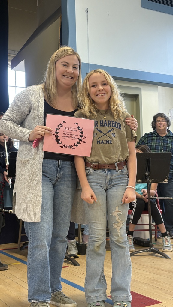 A teacher smiles next to a girl holding a pink award certificate; students sit in the background.