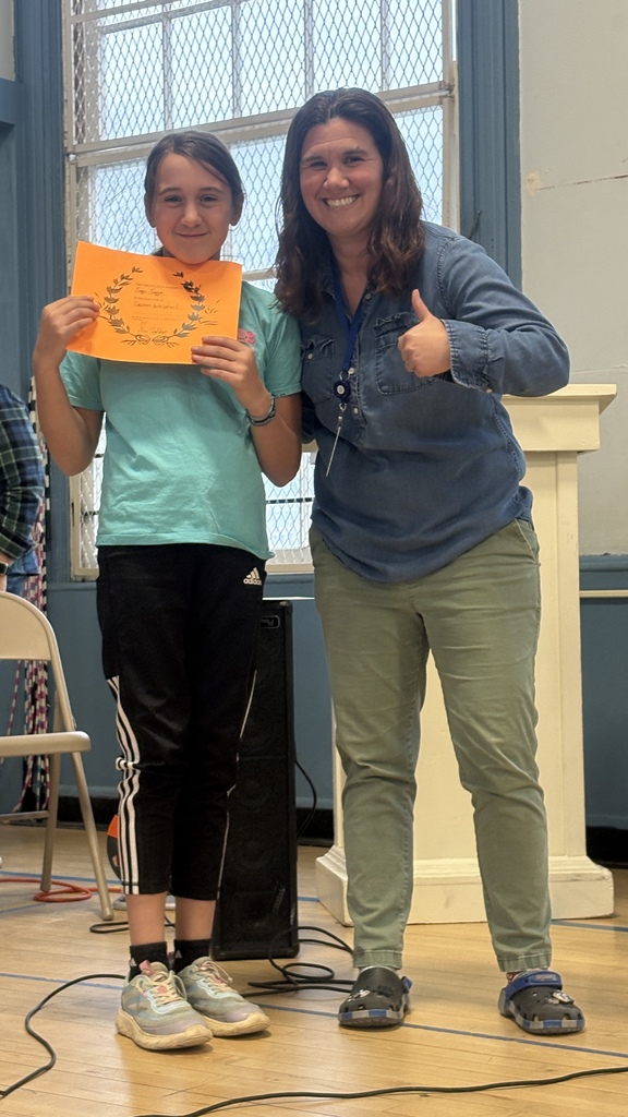 A teacher gives a thumbs-up while standing beside a girl holding an orange certificate.