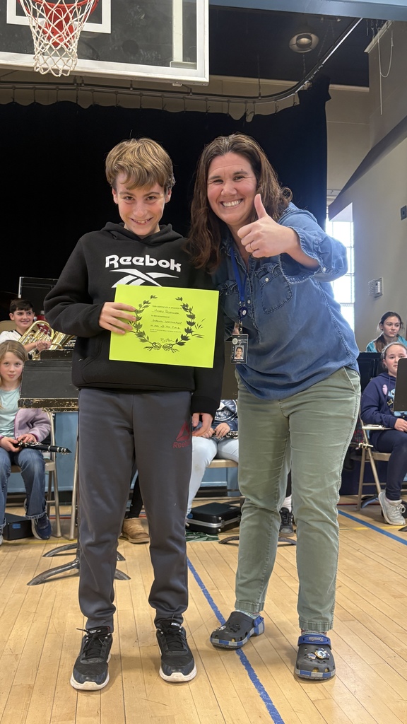 A teacher gives a thumbs-up while standing with a boy holding a bright green certificate in front of the school band.